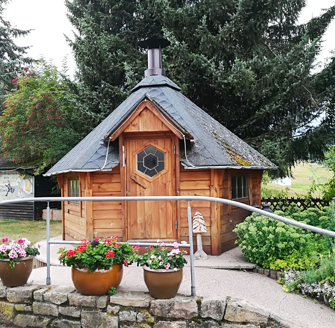 Kleine sechseckige Holzh&uuml;tte mit dunklem Dach und Schornstein, umgeben von Blumen und gr&uuml;nen Str&auml;uchern