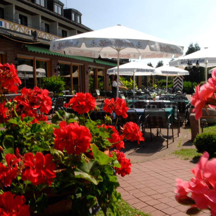 Au&szlig;enterrasse eines Hotel-Restaurants mit Tischen, St&uuml;hlen und Sonnenschirmen, umgeben von roten Blumen