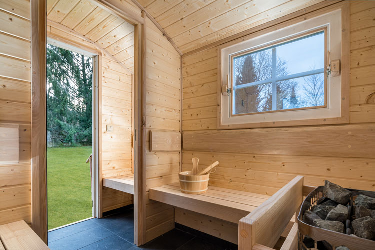 Innenansicht einer Holzsauna mit Bank, Aufgussk&uuml;bel und offenem T&uuml;rblick ins Gr&uuml;ne.