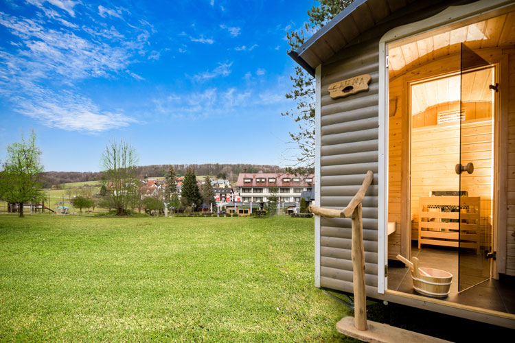Freistehende Au&szlig;ensauna mit Holzinterieur auf gr&uuml;ner Wiese vor Hotelanlage
