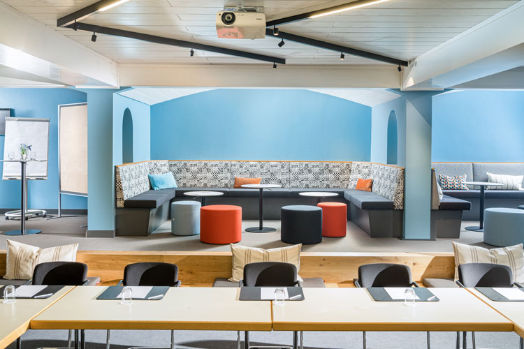 Moderner Seminarraum mit blauen W&auml;nden, gepolsterter Sitzecke, Hockern und Tischen f&uuml;r Gruppenarbeit