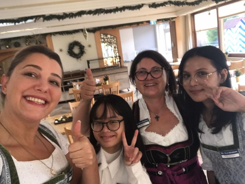 Vier Personen in Dirndl und Trachten posieren in einem festlich dekorierten Raum mit Holzst&uuml;hlen.