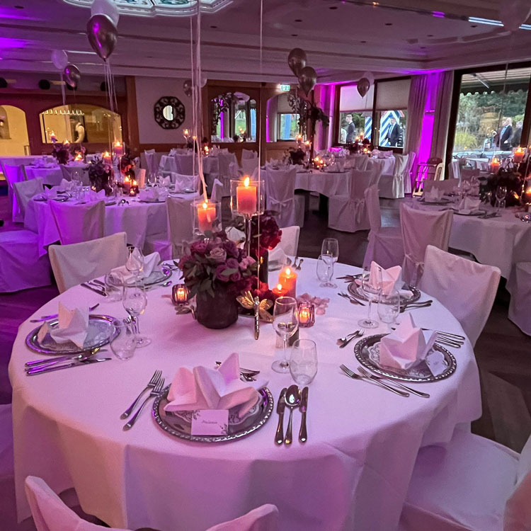 Festlich dekorierter Bankettsaal mit runden Tischen, wei&szlig;en Hussen, Kerzen, Blumen und violetter Beleuchtung