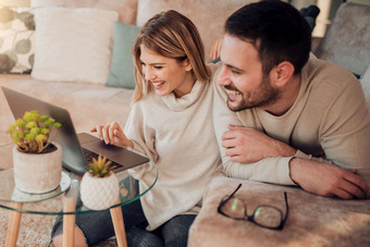 Zwei lächelnde Personen sitzen auf Sofa und schauen auf einen Laptop auf Glastisch