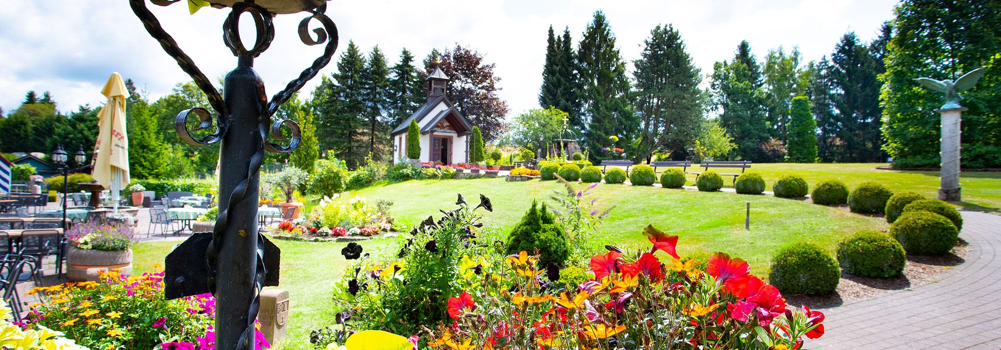 Blühender Hotelgarten mit Sitzbereich, Blumenbeeten und kleiner Kapelle im Hintergrund
