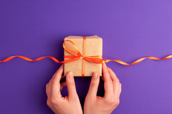 Geschenk in braunem Papier mit orangefarbener Schleife, gehalten von zwei Händen vor violettem Hintergrund