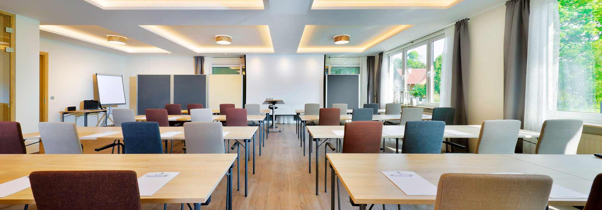 Heller Seminarraum mit rechteckigen Tischen in Reihen, Stühlen, Flipchart und großer Projektionsfläche