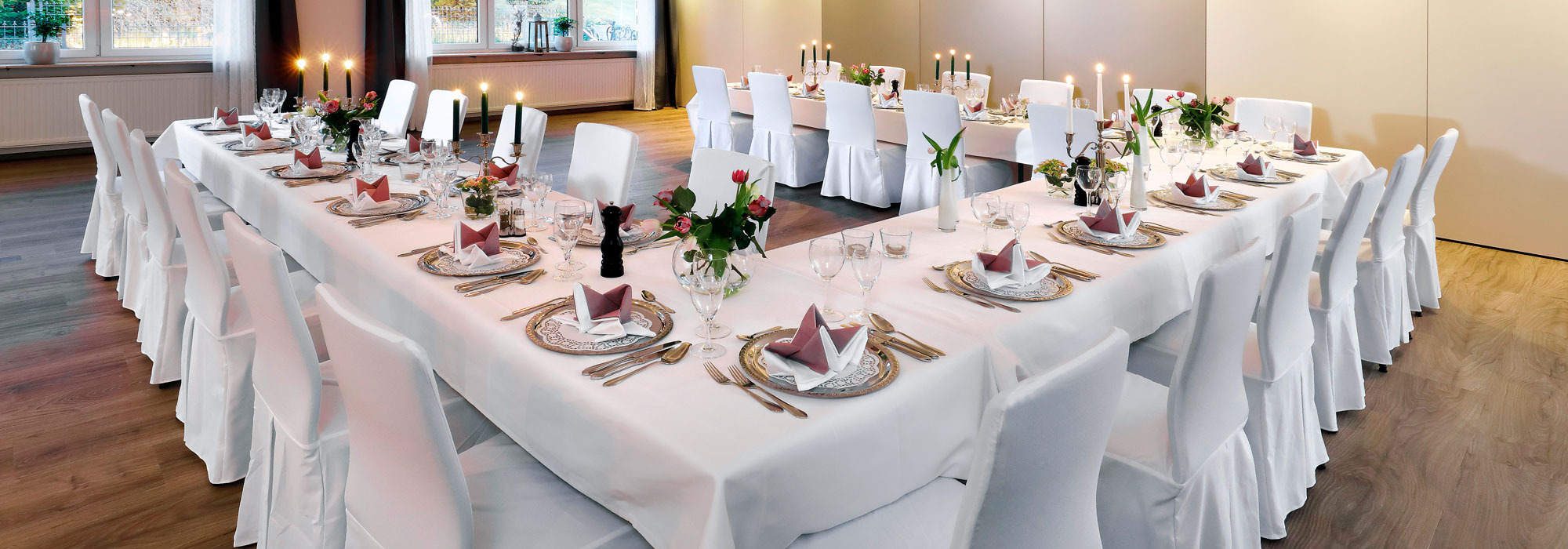 Festlich gedeckter U-förmiger Tisch mit weißen Hussen, Kerzen und Blumenarrangements in elegantem Raum