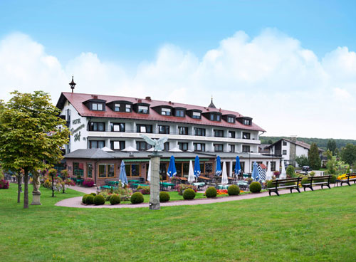 Hotel mit Au&szlig;ensitzbereich, umgeben von B&auml;umen und Gr&uuml;n unter blauem Himmel.